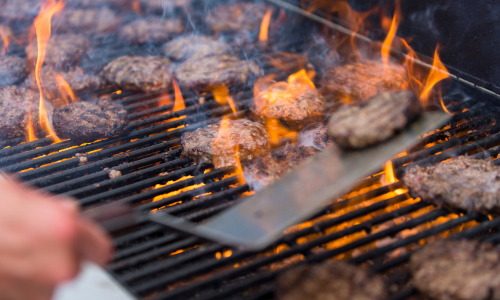Hamburgers on a propane grill, being cooked and flipped with a spatula.